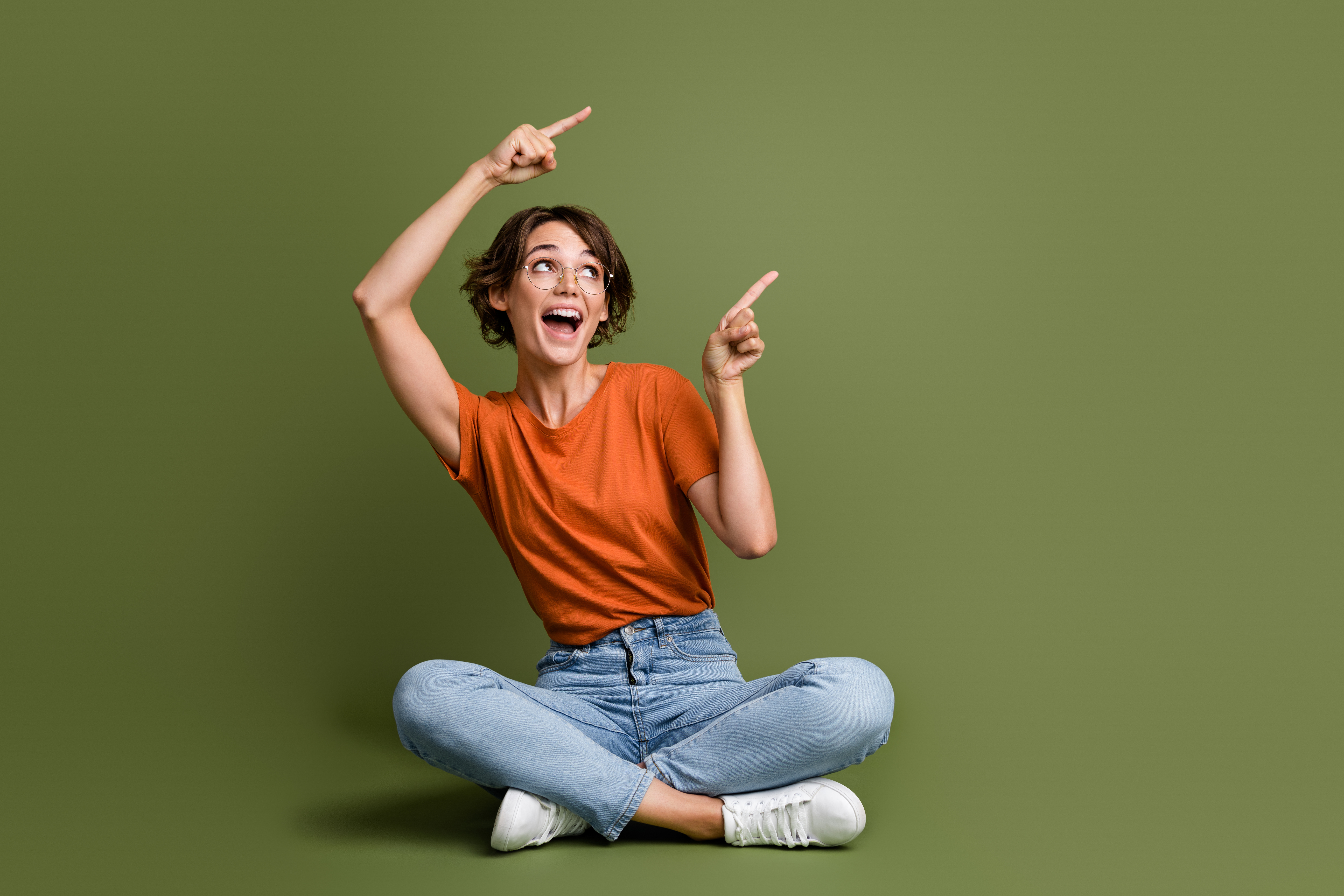 Full length photo of impressed girl dressed t-shirt sit on floor look directing at promo empty space isolated on green color background.